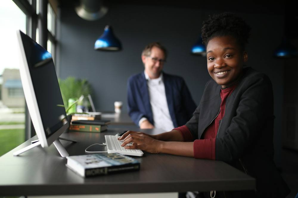 A smiling professional at her desk in a modern office, with a colleague working behind her