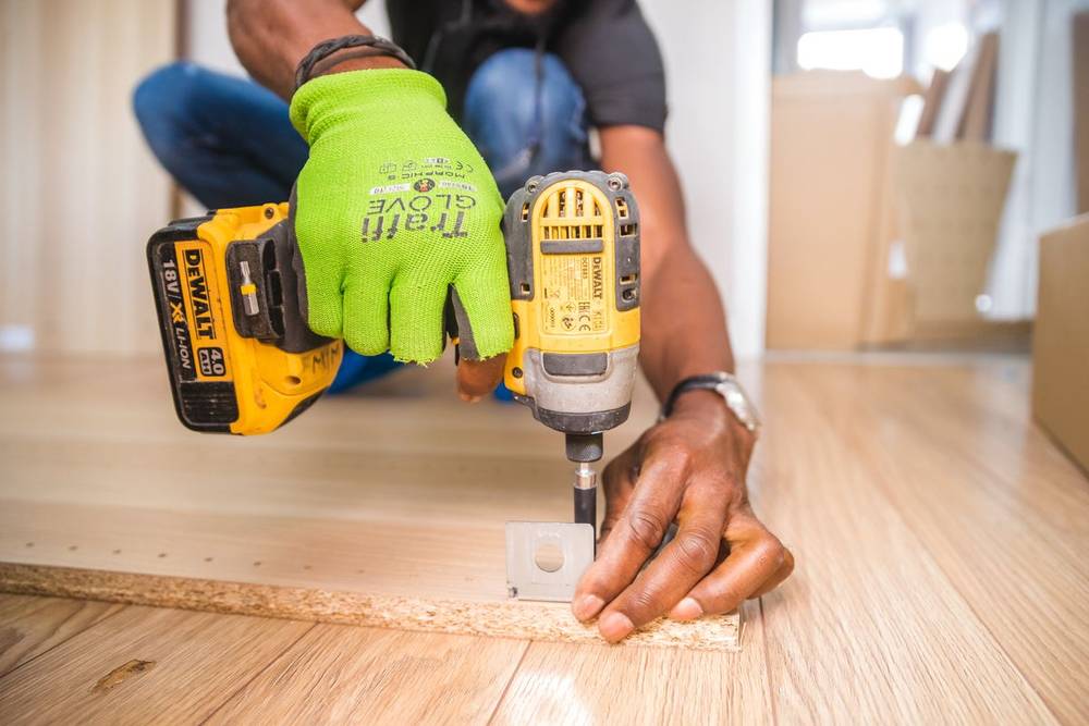 A tradesperson in bright work gloves driving a screw into a timber floor with a cordless drill
